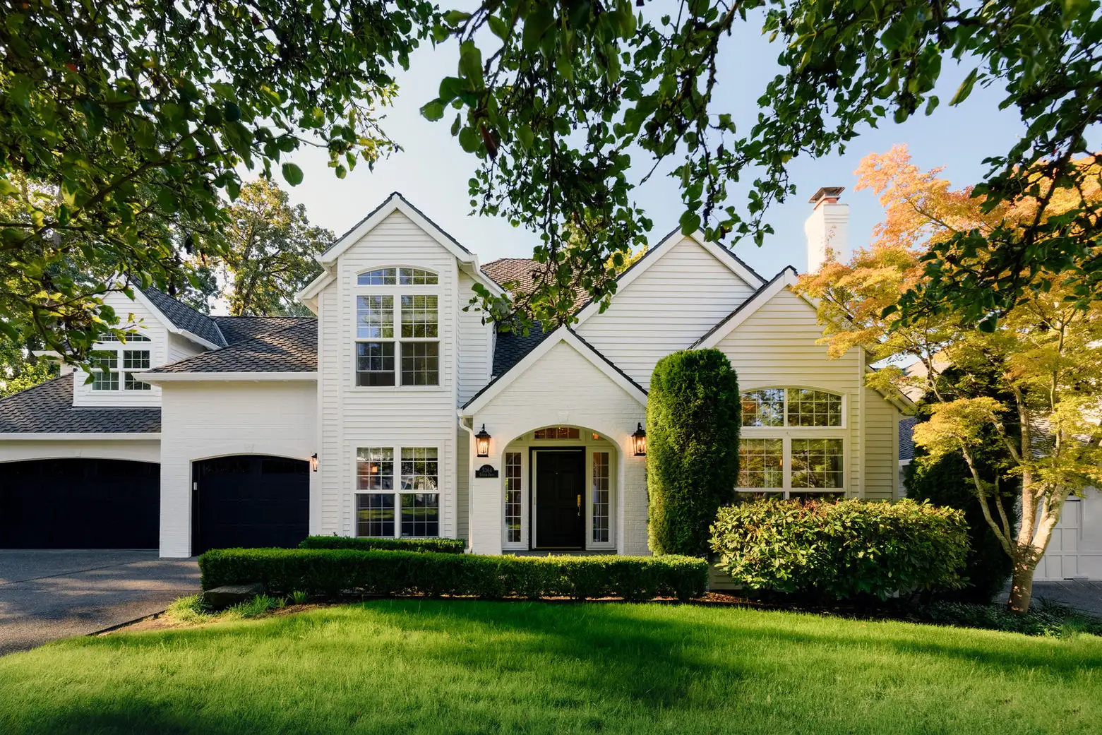 White two-story remodeled home with steep gables, a black front door, and manicured hedges
