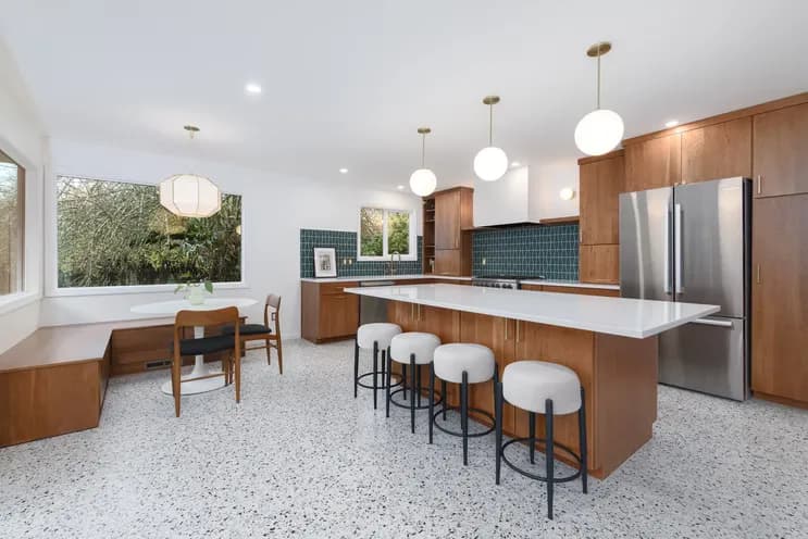After photo of a renovated SE Portland kitchen showing a large white quartz island with boucle stools, built-in walnut banquette dining nook, picture window with garden views, teal tile backsplash, and terrazzo floors by Tortoise Construction LLC