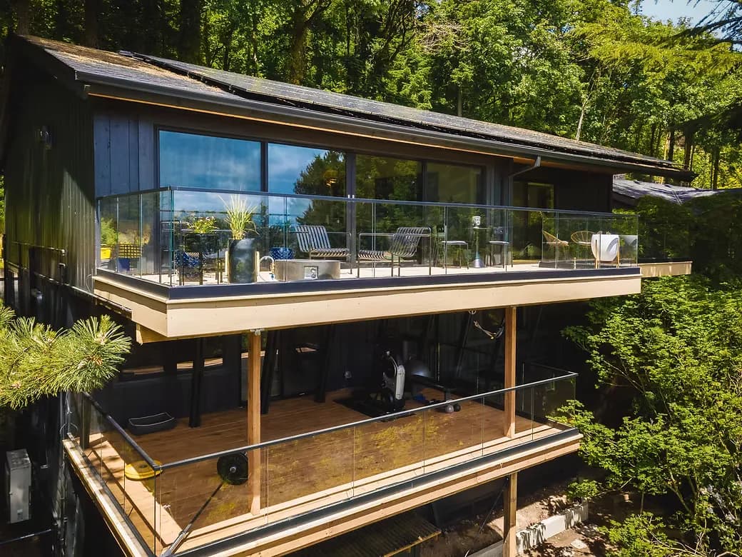 Exterior view of a two-story modern home surrounded by tall trees, featuring dual-level decks with frameless glass panel railings, solar panels on a dark roof, floor-to-ceiling windows, and outdoor furniture on the upper deck