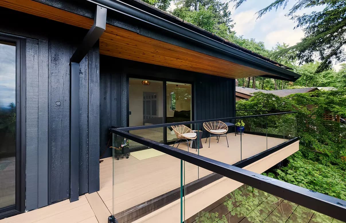 Elevated private balcony with light composite decking, black-framed frameless glass railing, and two wicker chairs, accessed by sliding glass doors from a bedroom on a dark navy board-and-batten home surrounded by lush green trees