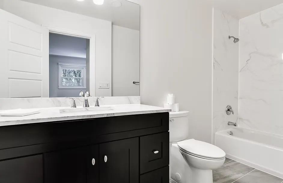 Remodeled guest bathroom with a dark shaker-style vanity, white marble countertop, chrome faucet, large frameless mirror, white toilet, marble-surround tub-shower combo with chrome fixtures, and wood-look gray tile flooring against bright white walls.