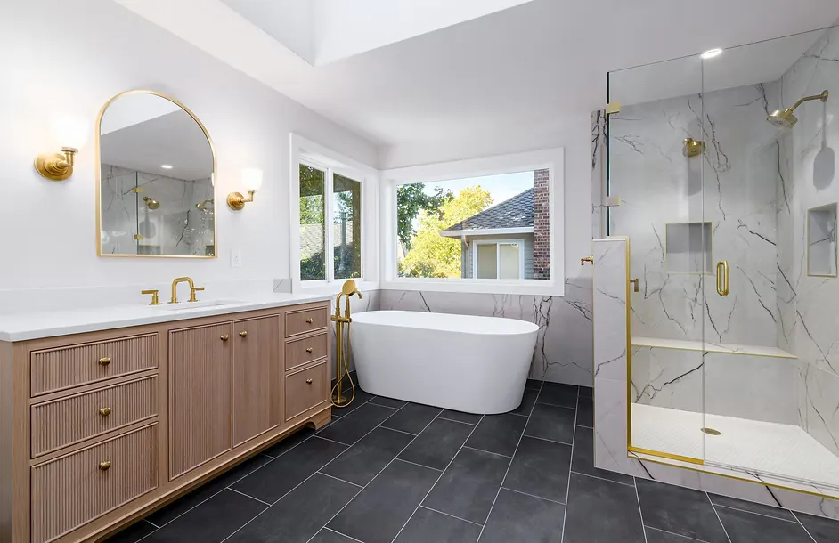 Remodeled master bathroom from the vanity-side angle, featuring a fluted natural wood single vanity with brushed gold hardware and white countertop, arched gold-framed mirror with flanking gold wall sconces, a white oval freestanding soaking tub beneath a large picture window, a gold-framed frameless glass walk-in shower with floor-to-ceiling white marble tile, built-in shower niche, bench, and multiple gold fixtures, and dark large-format slate floor tiles.