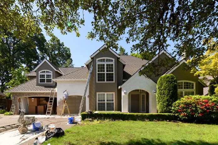 Exterior of a Lake Oswego home mid-renovation with a worker on a ladder painting the front facade, construction materials on the driveway, and mature trees framing the property — Tortoise Construction LLC full home remodel in progress.