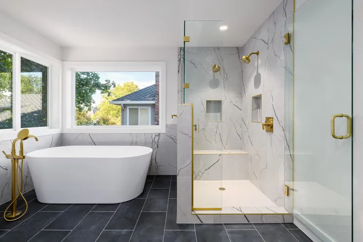 Remodeled bathroom with a freestanding white soaking tub, brass floor-mounted tub filler, marble-look shower surround behind a glass enclosure with brass fittings, and large dark slate floor tiles