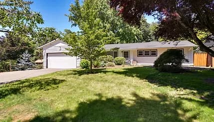 Single-storey ranch home with light siding and an attached two-car garage, set back behind a mown lawn and mature shade trees
