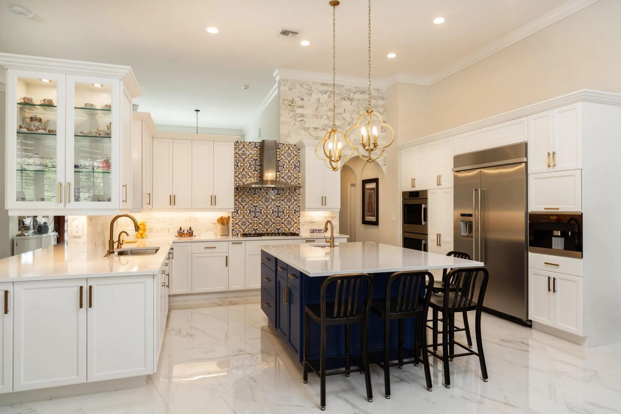 Open-plan kitchen with white shaker cabinetry, a navy blue island seating four, brass quatrefoil pendant lights, patterned blue and gold tile behind the range, and polished marble floors