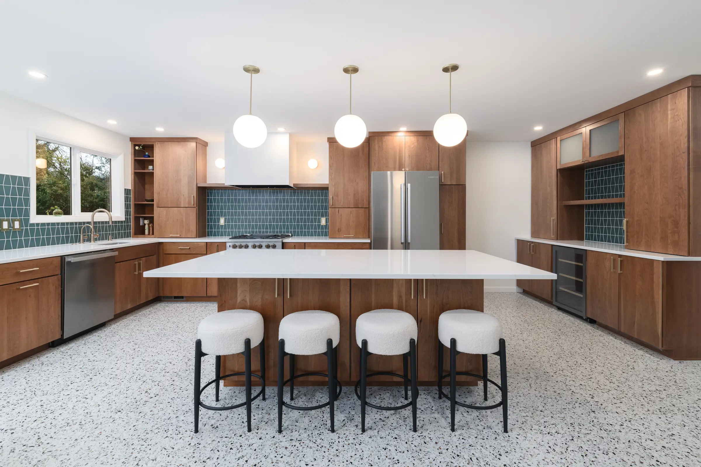 After photo of a fully renovated SE Portland kitchen featuring custom walnut cabinetry, a large white quartz island with four boucle stools, teal geometric tile backsplash, three brass globe pendants, and terrazzo floors by Tortoise Construction LLC